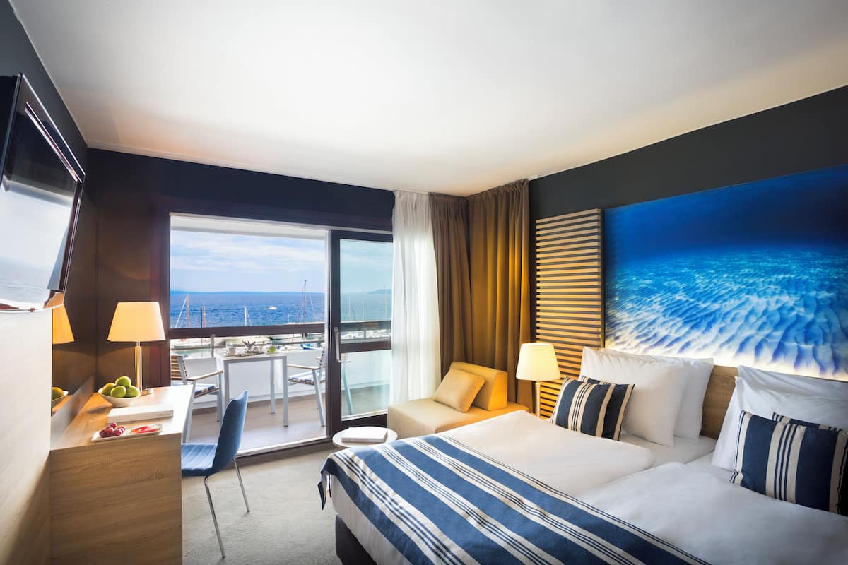 Een hotelkamer met een groot bed, een bureau met lamp, een stoel, een klein tafeltje met fruit, en uitzicht op de oceaan door een groot raam.