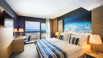 A hotel room with a large bed, a desk, a chair, a TV, and a view of the ocean.