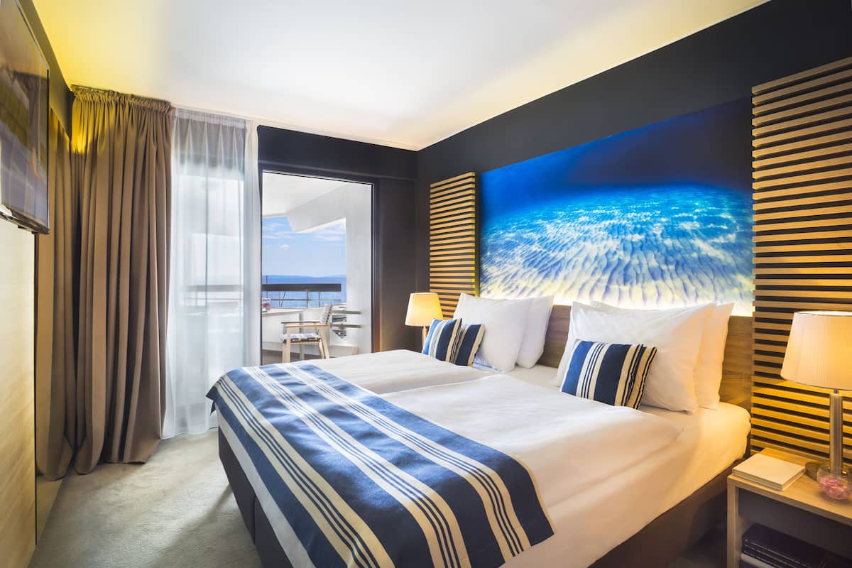 Een hotelkamer met een groot bed, uitzicht op zee, een balkon en een bureau met een lamp.