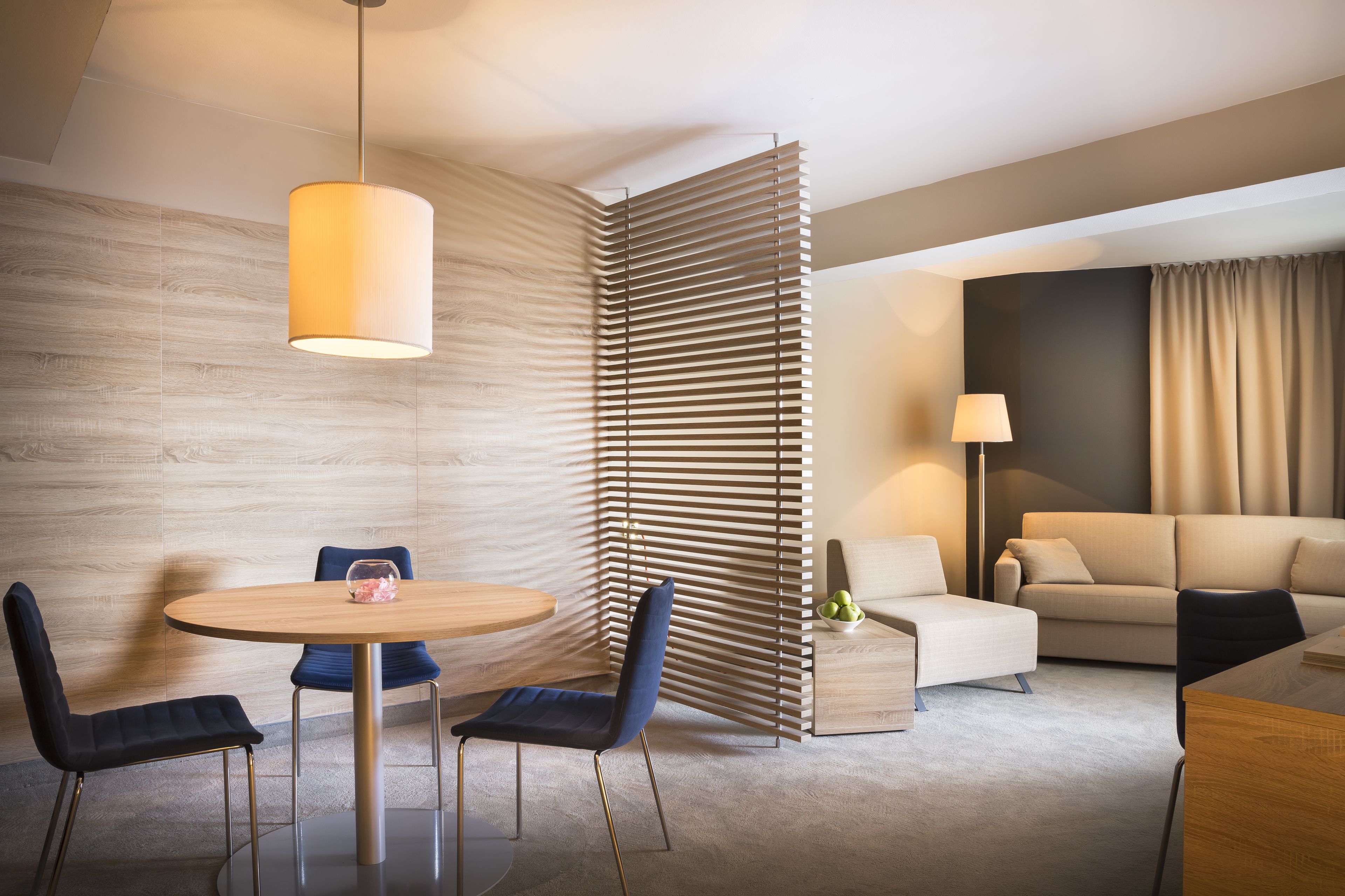 A modern hotel room with a round dining table, two chairs, a sofa, a lamp, and a decorative partition.