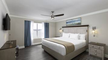 A hotel room with a large bed, a desk, a chair, a ceiling fan, a TV, and a window with curtains.