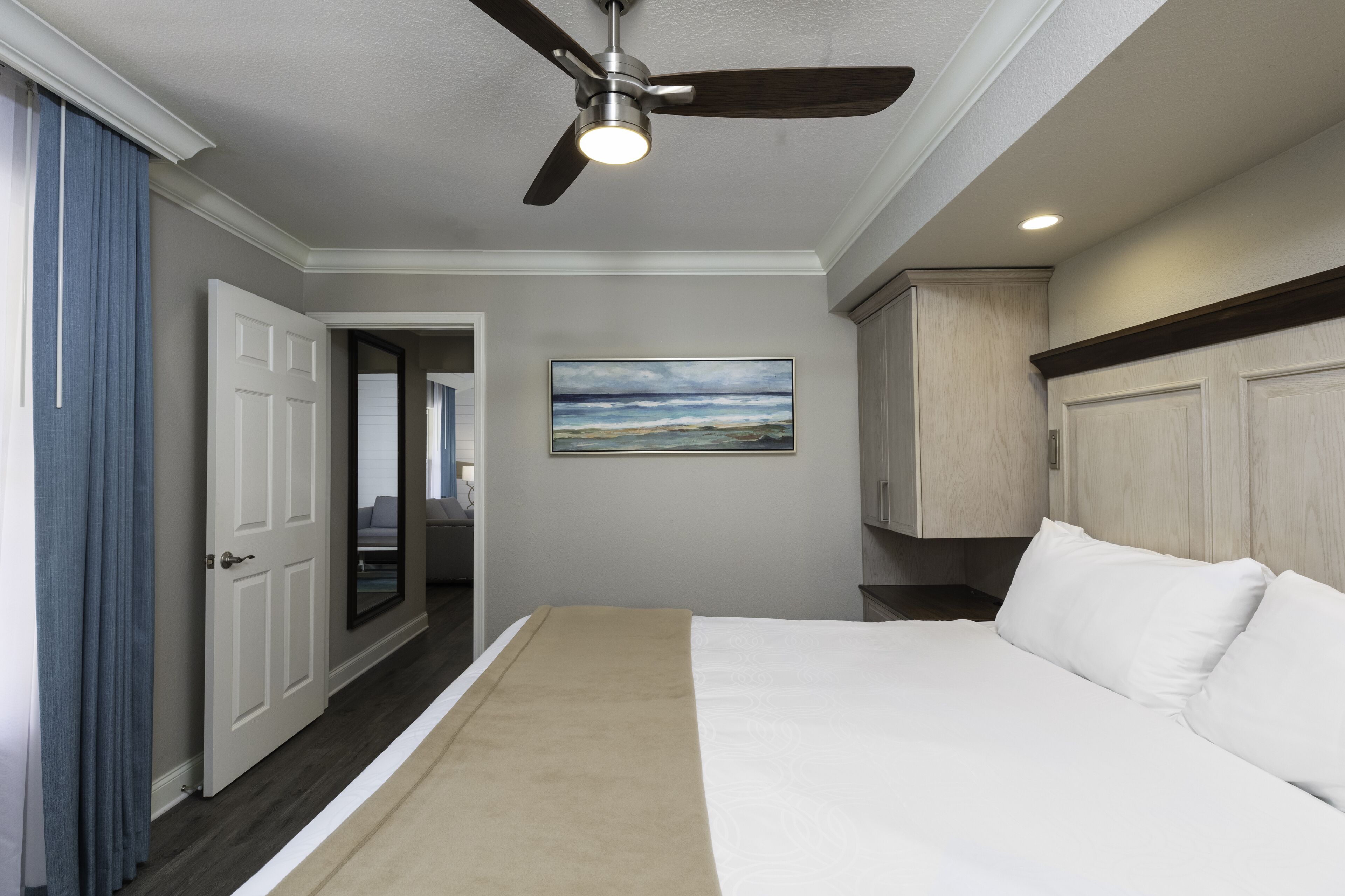 A hotel room with a bed, a ceiling fan, a painting on the wall, and a door leading to another room.
