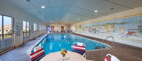 An indoor swimming pool with a mural, a table with a vase of flowers, and striped chairs.