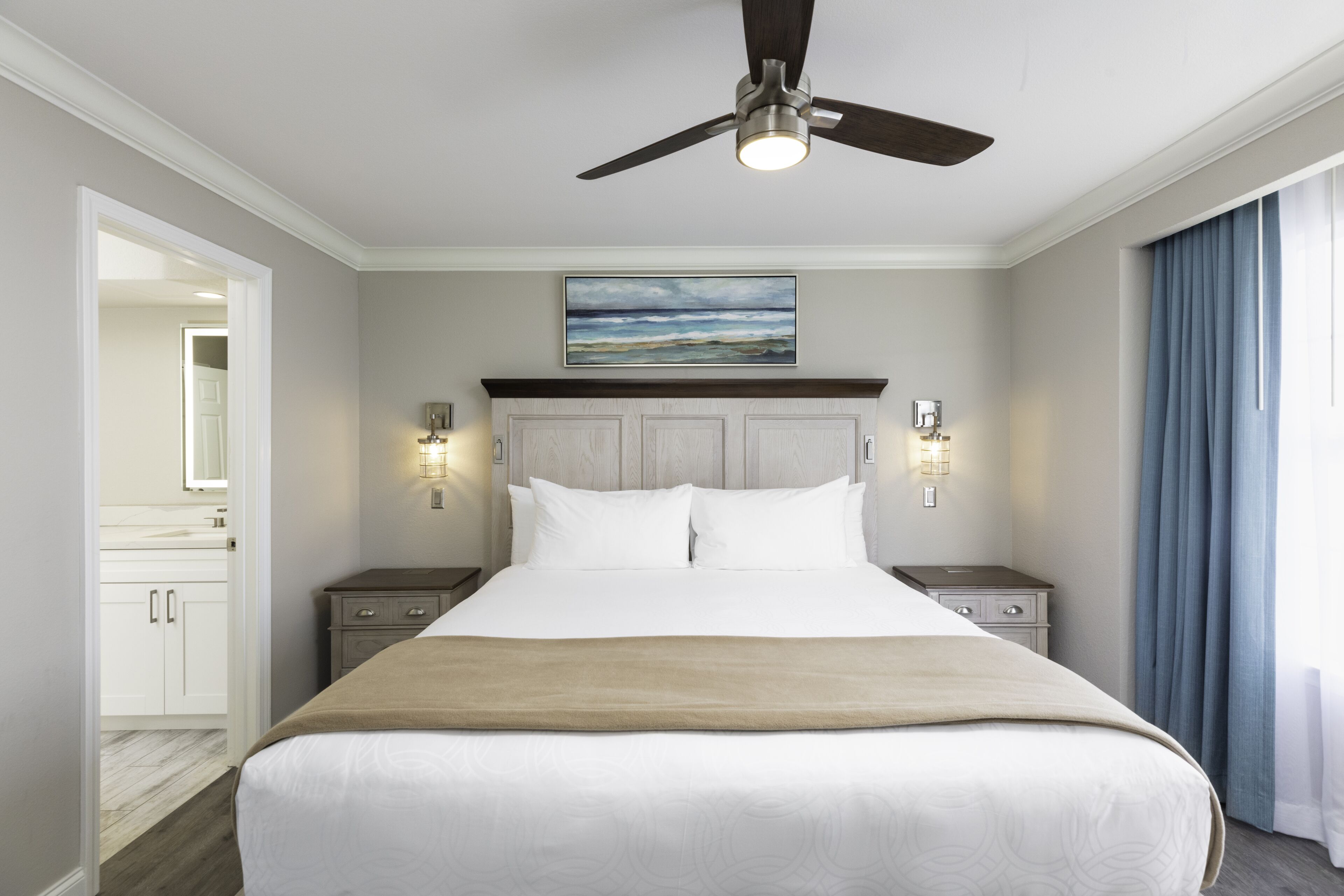 A hotel room with a large bed, a ceiling fan, a painting above the bed, and a view of the ocean through the window.