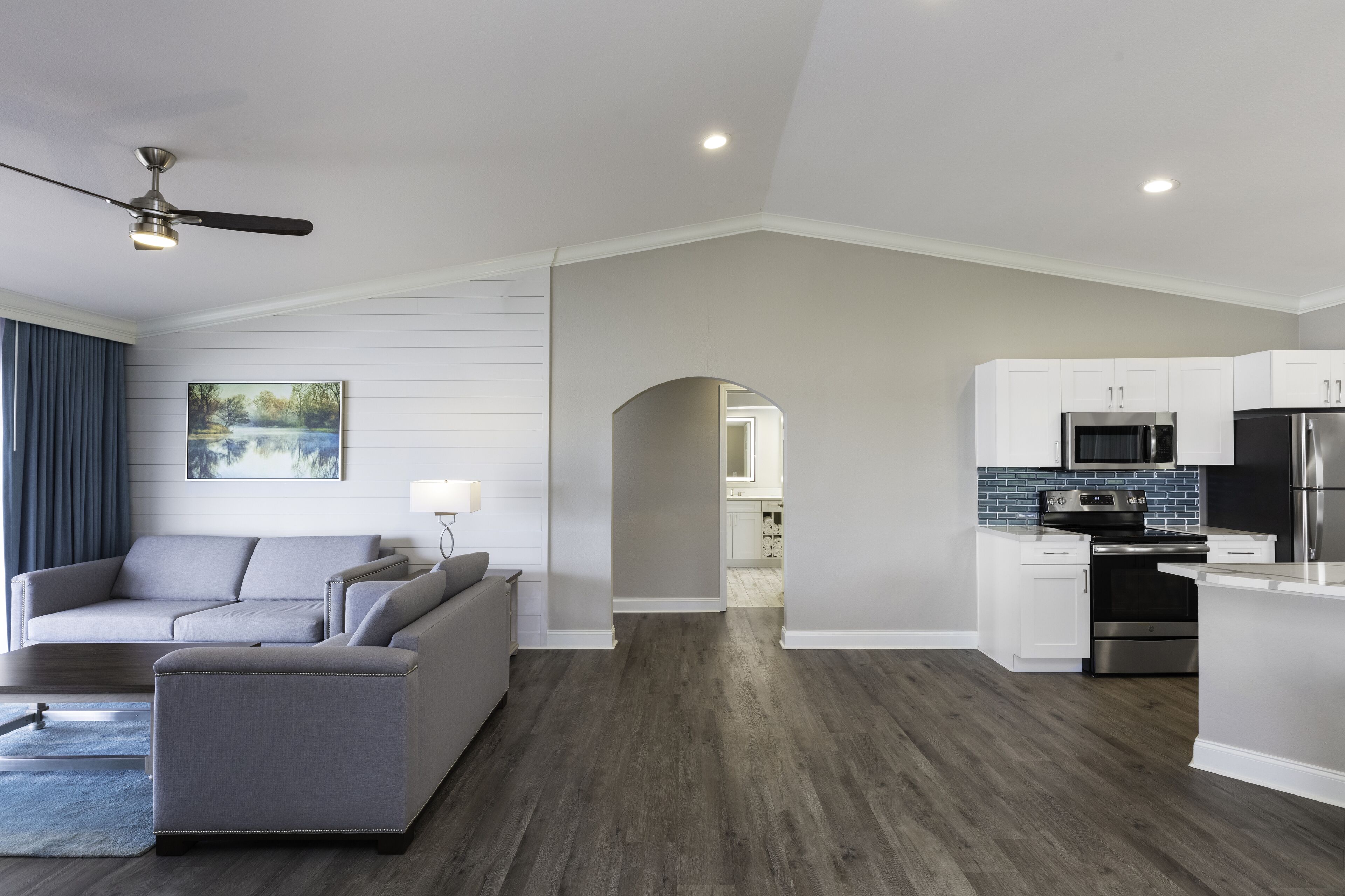 A modern living room with a grey sofa, a small ottoman, and a ceiling fan. A kitchen with white cabinets and stainless steel appliances is visible in the background.