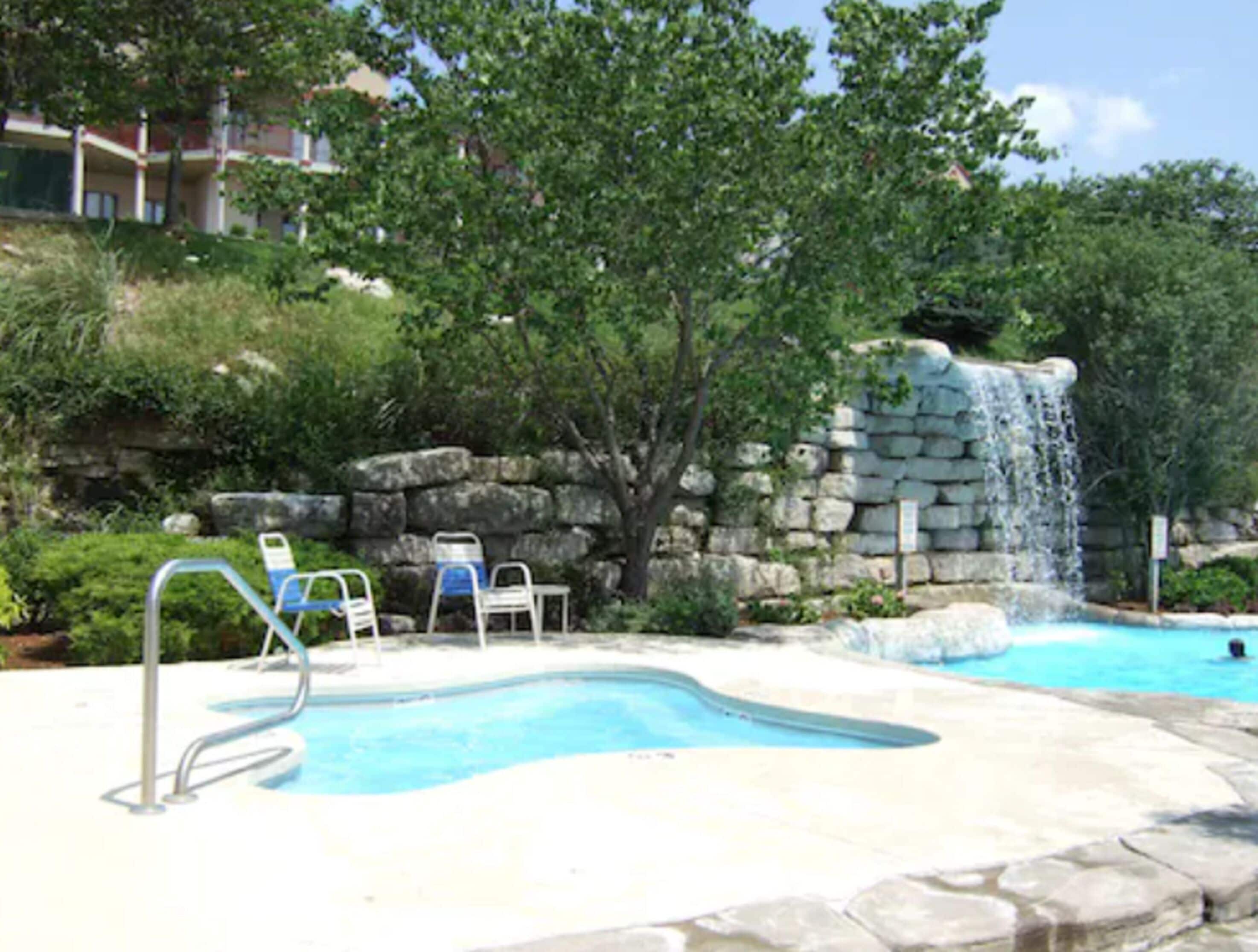 A swimming pool with a stone waterfall feature, surrounded by greenery and a stone wall.