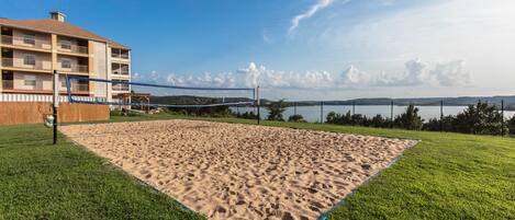 A sandy volleyball court with a net, surrounded by green grass, and a building with multiple balconies in the background.