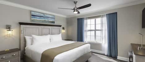 A hotel room with a large bed, a ceiling fan, a nightstand, a desk, and a window with curtains.