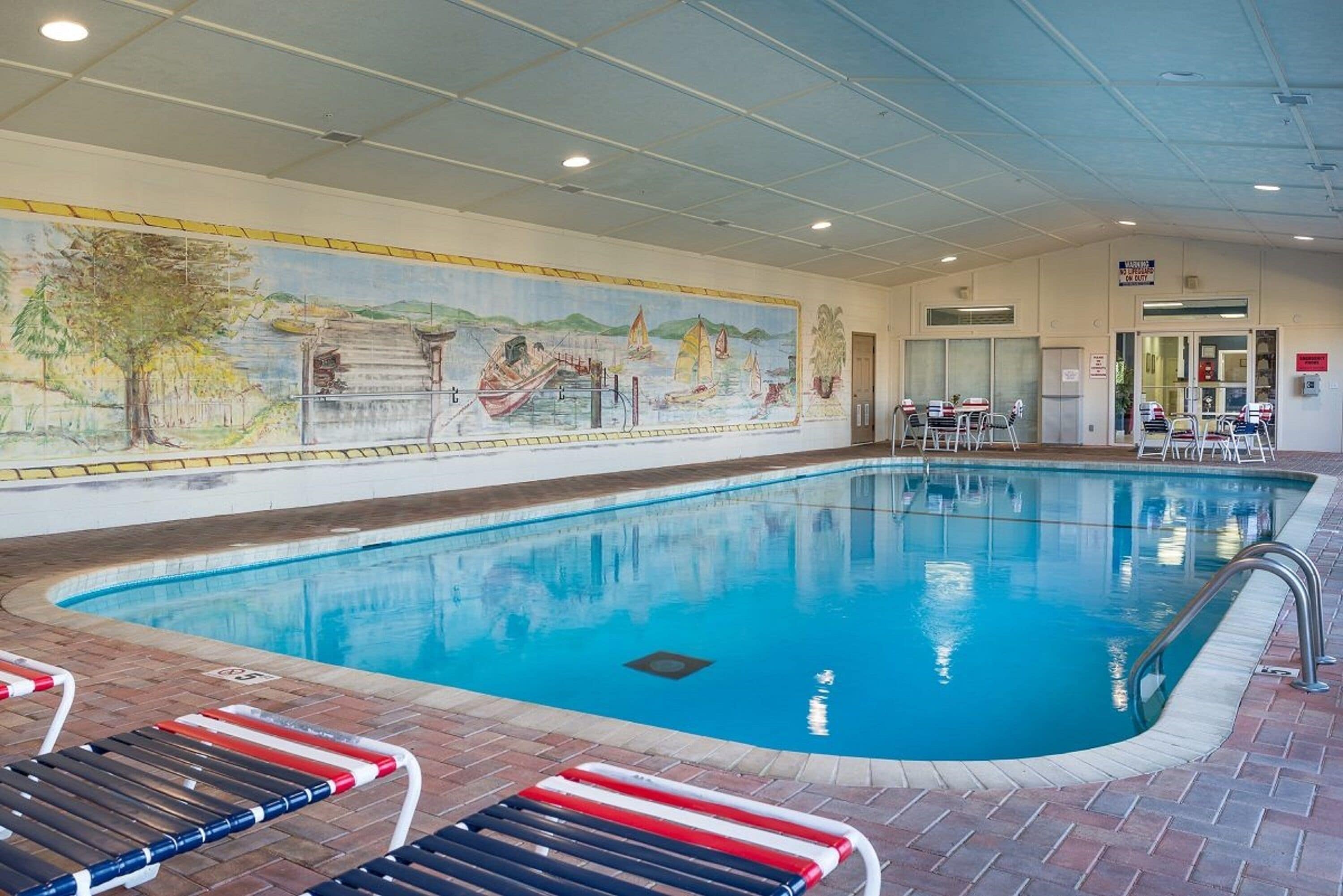 Une piscine intérieure ornée d’une fresque murale, avec des chaises longues et un espace d’accueil.