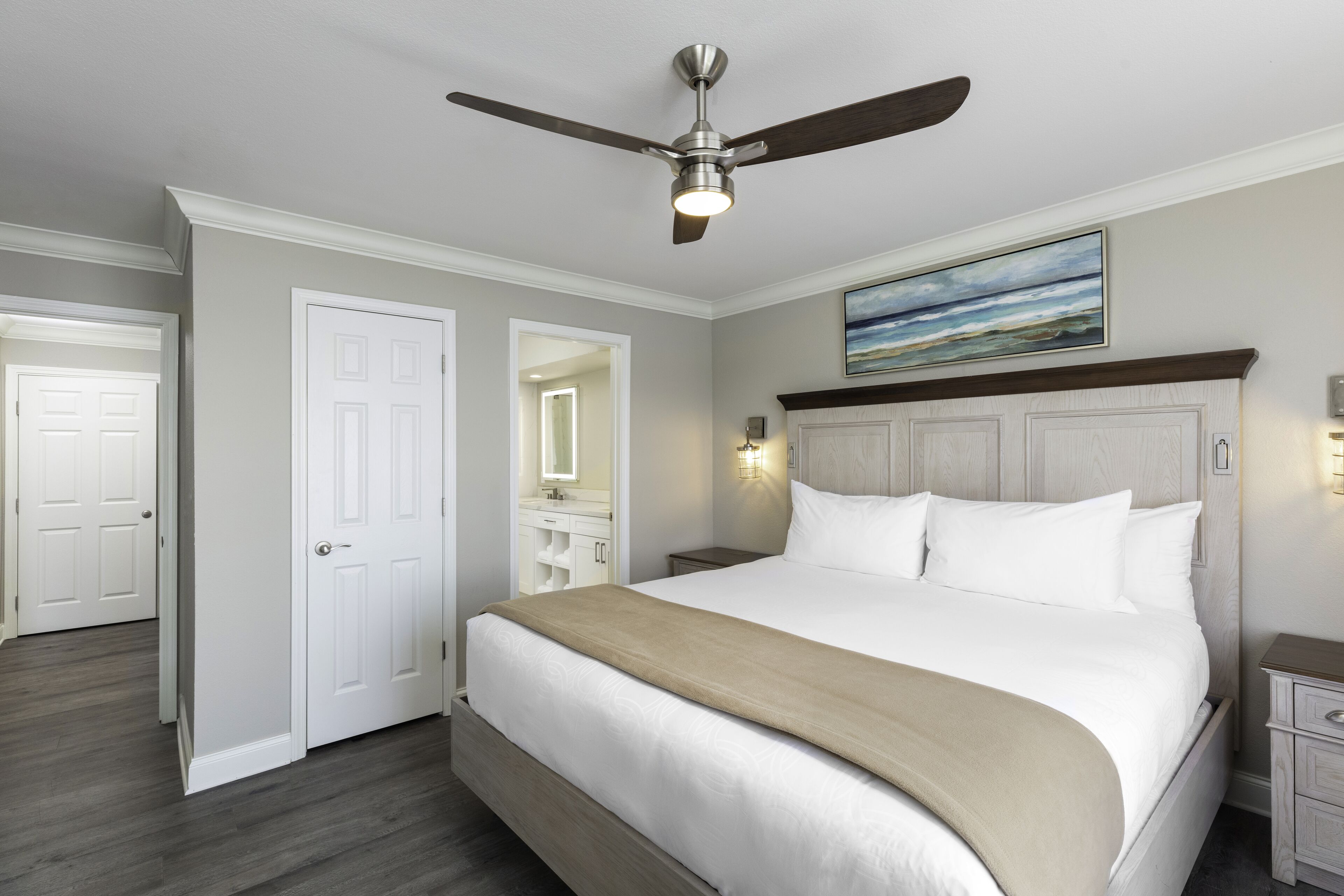 A hotel room with a large bed, a ceiling fan, a painting on the wall, and a wooden floor.