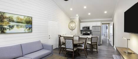 A modern kitchen with white cabinetry, a dining area with a round table and chairs, a gray sofa, and a ceiling fan.