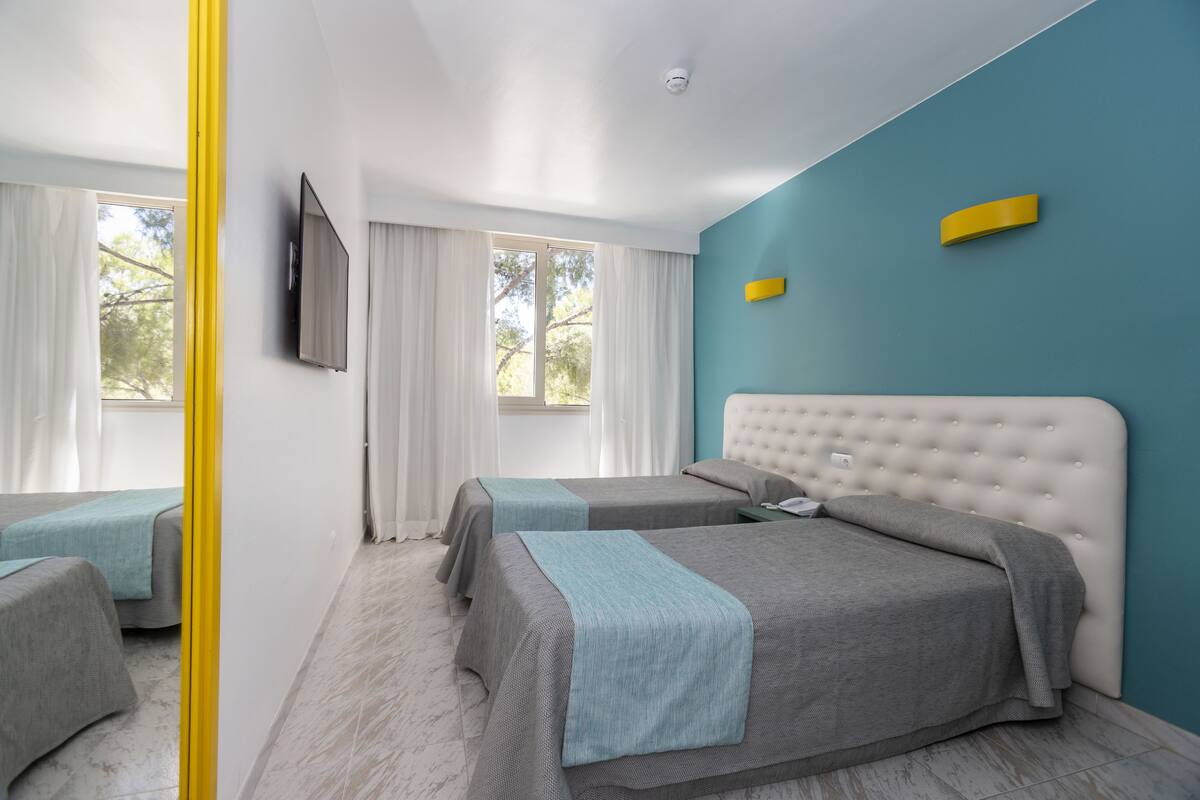 A modern hotel room with two beds, a large window, and a teal accent wall.