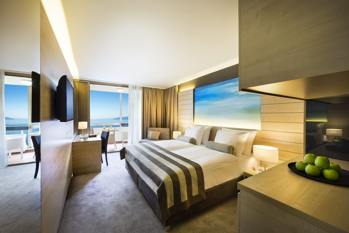 A hotel room with a large bed, a desk, a chair, a nightstand, a lamp, and a view of the ocean.