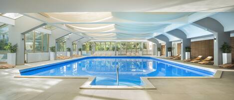 A large indoor swimming pool with clear blue water, surrounded by sun loungers and potted plants.