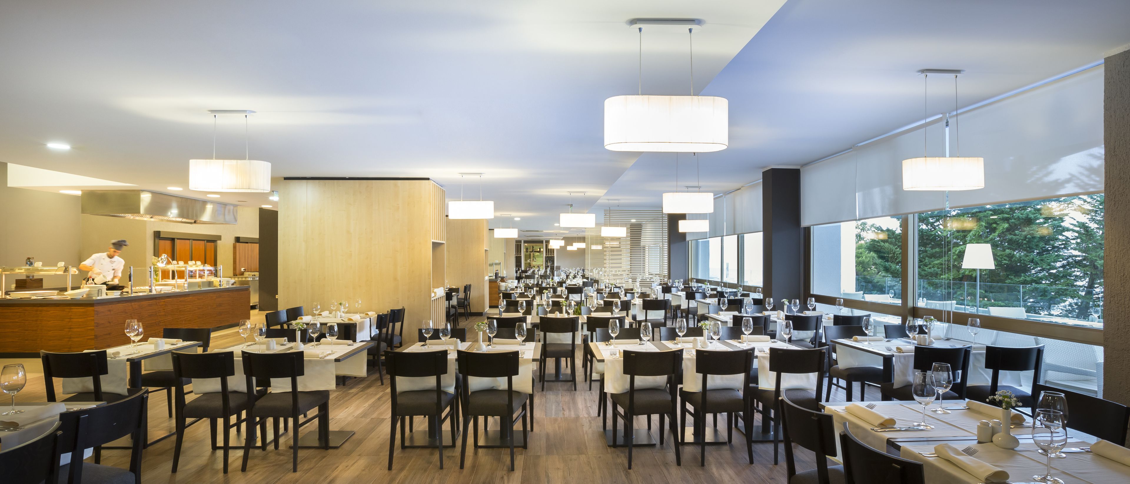A spacious restaurant with multiple dining tables and chairs, large windows, and modern pendant lighting.