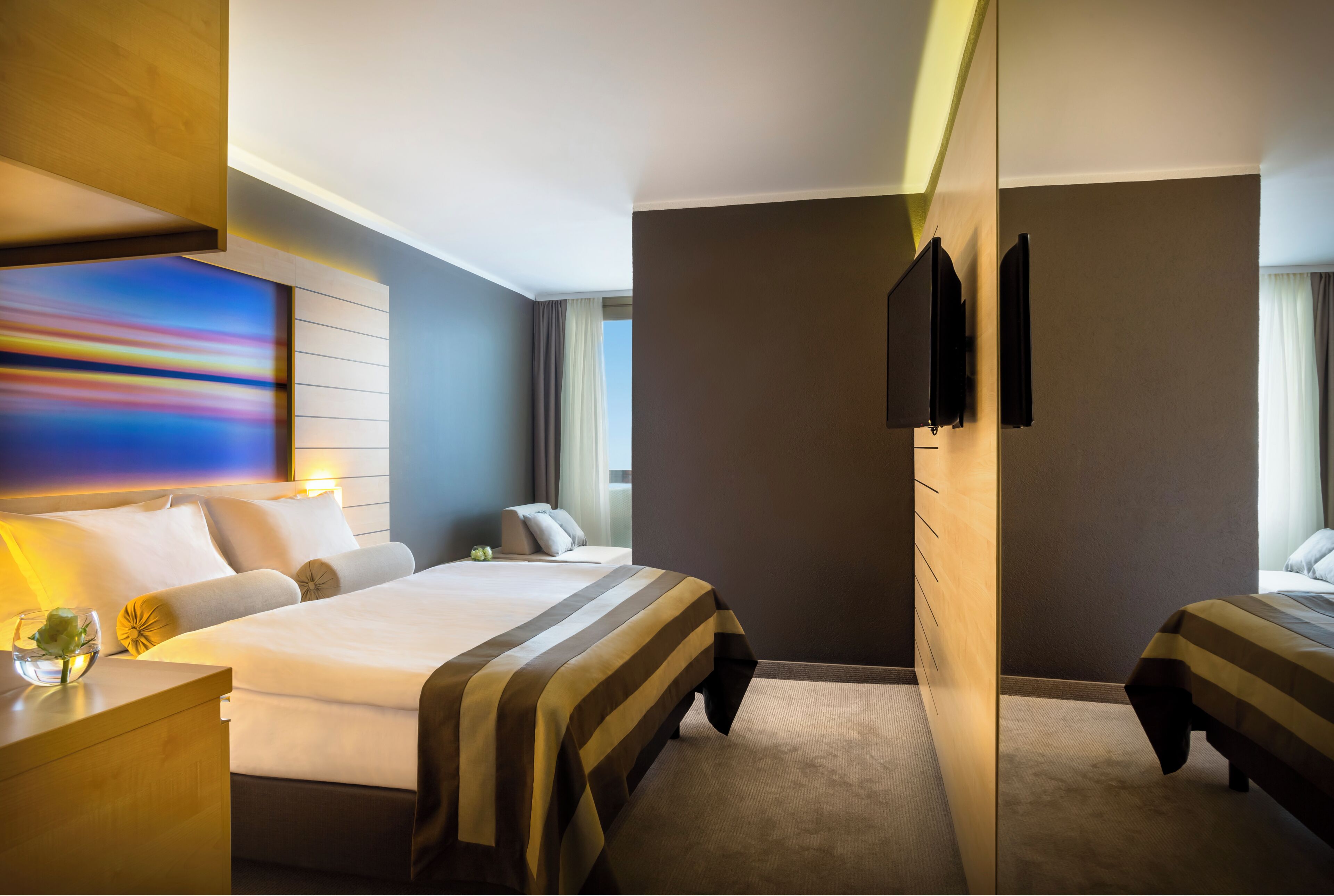 A hotel room with a large bed, a bedside table with a lamp, a TV mounted on the wall, and a window with curtains.