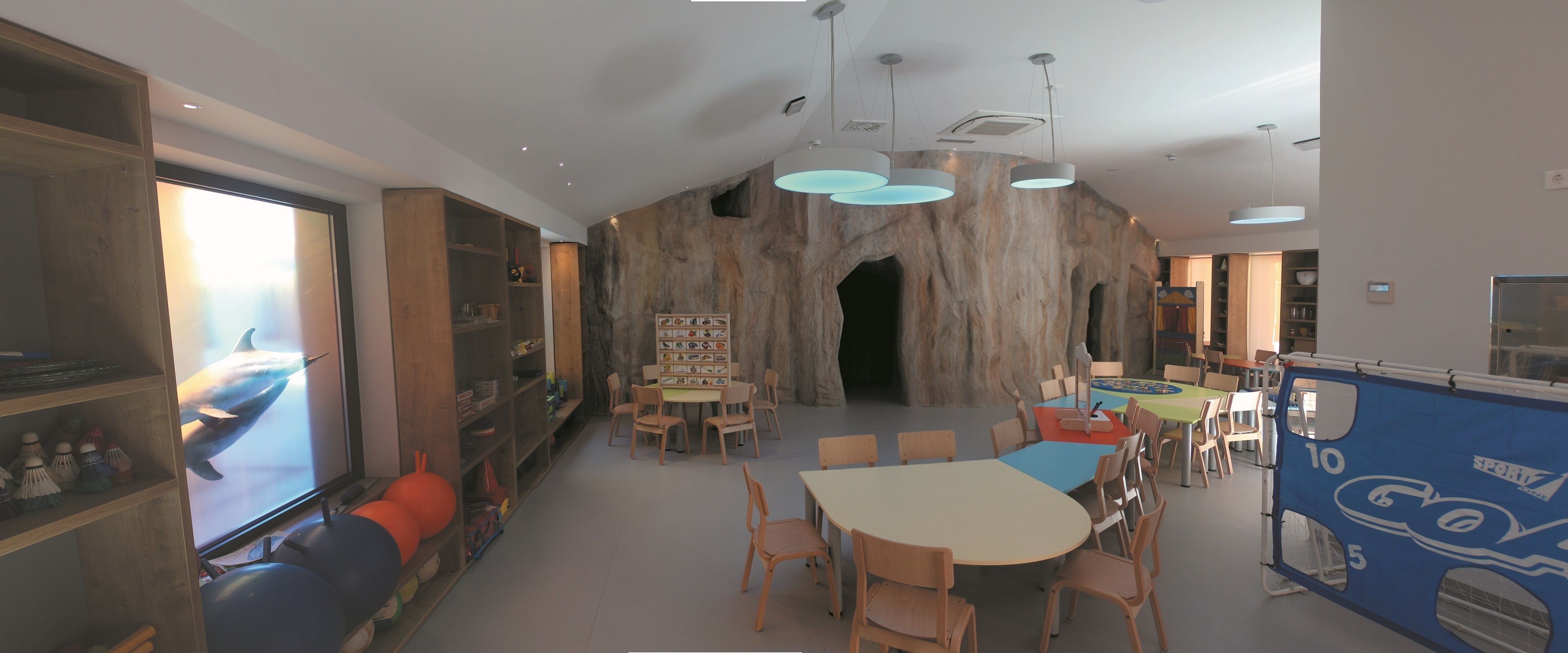 A spacious room with a large table and chairs, a bookshelf, and a wall with a rock-like texture.