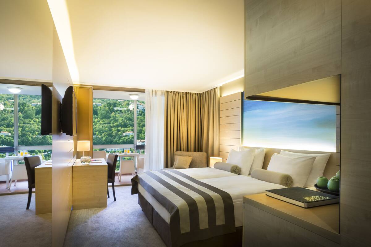 A hotel room with a large bed, a desk, and a chair. There is a TV mounted on the wall and a window with curtains.