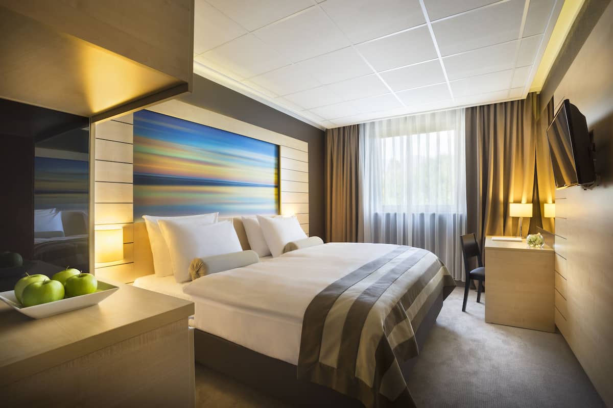 A hotel room with a large bed, a bedside table with a bowl of apples, a desk with a lamp, a flat-screen TV, and a painting on the wall.
