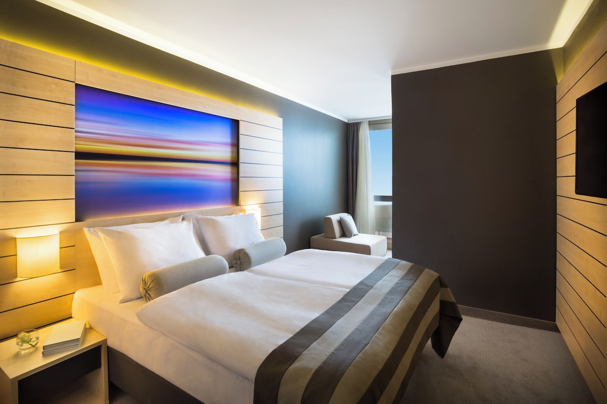 A modern hotel room with a large bed, bedside tables, a view of the city, and a large abstract painting on the wall.
