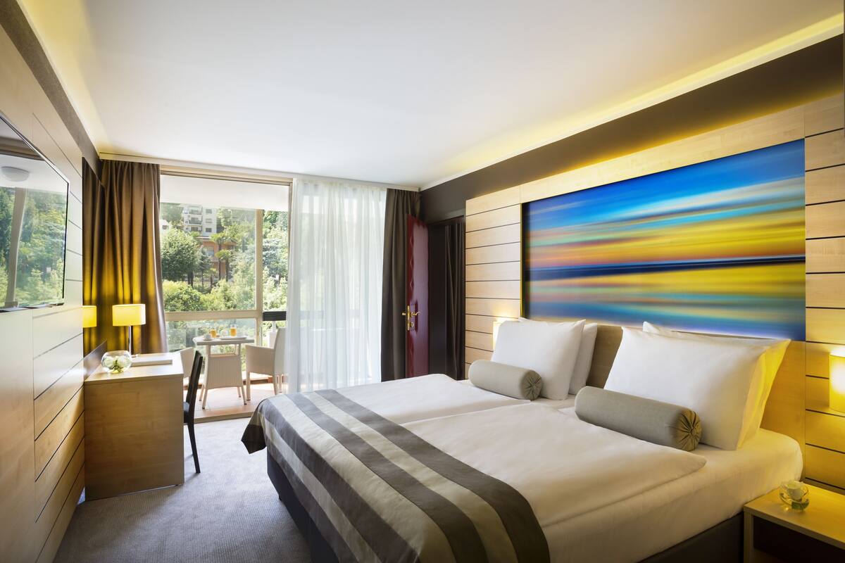 A hotel room with a large bed, a desk with a lamp, a chair, and a view of greenery through the window.