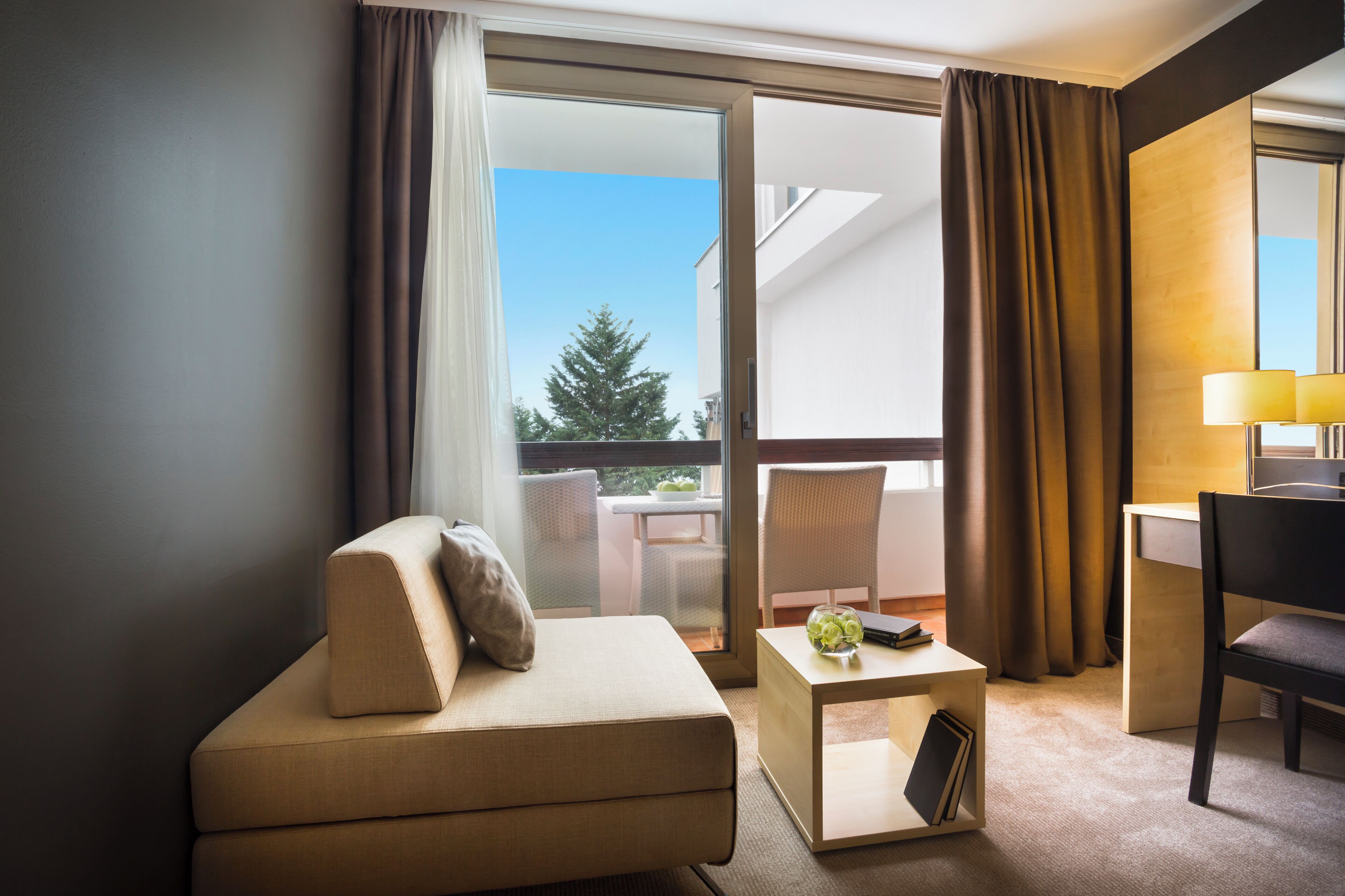 Une chambre d’hôtel avec un canapé, une petite table avec une lampe, une chaise et une vue sur un balcon agrémenté d’un arbre.