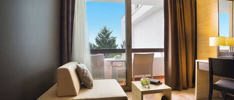 Une chambre d’hôtel avec un canapé, une petite table avec une lampe, une chaise et une vue sur un balcon agrémenté d’un arbre.