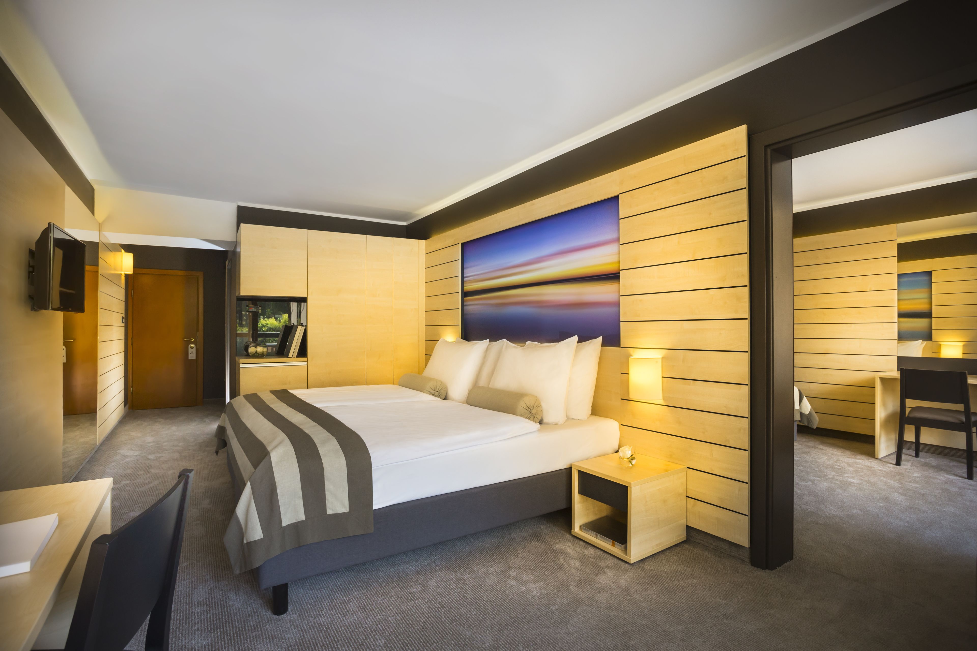 A modern hotel room with a large bed, bedside tables, a desk, and a painting on the wall.