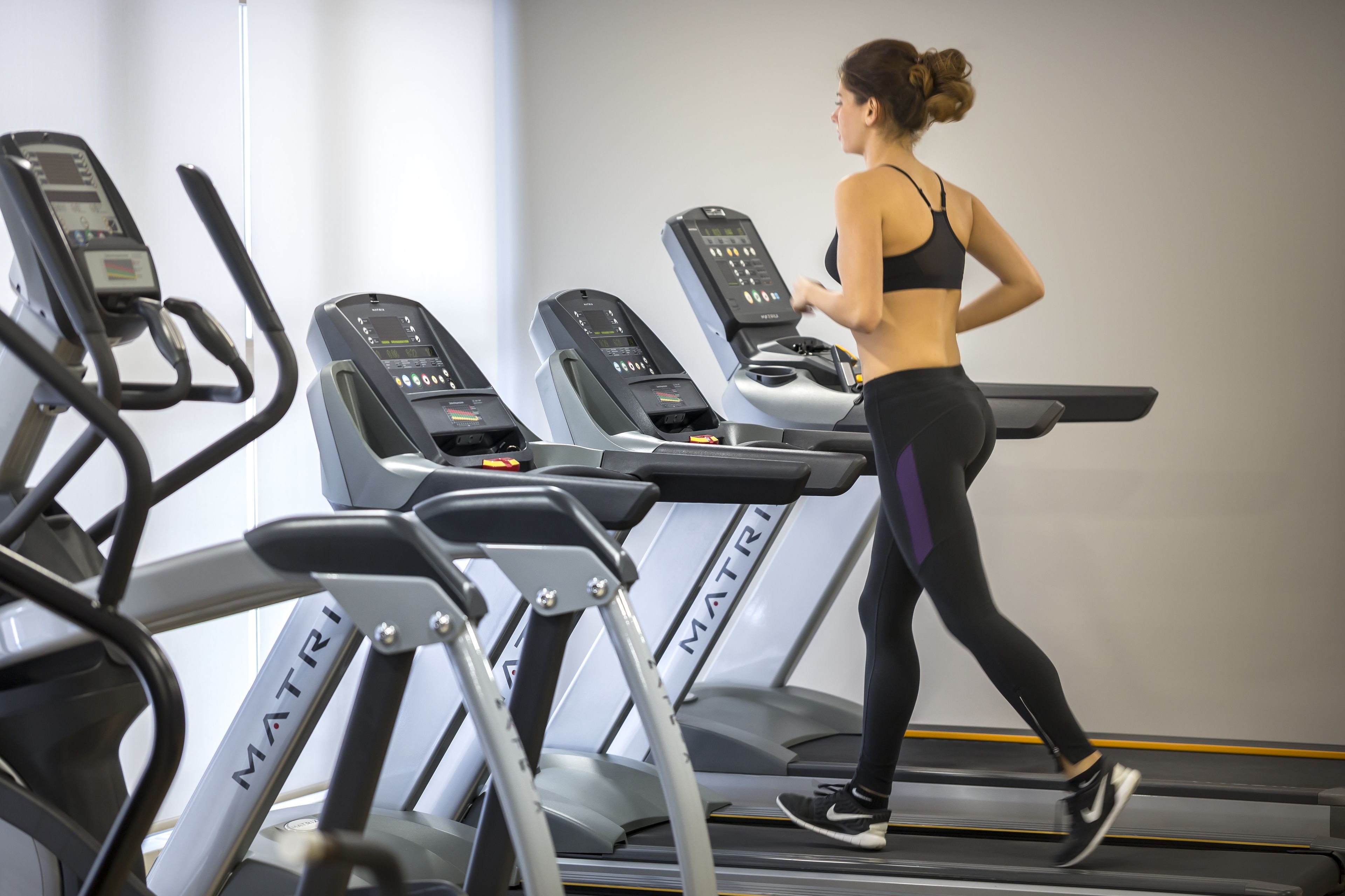 Une femme court sur un tapis de course dans une salle de sport où plusieurs tapis de course sont disposés et portent l'inscription « MATRIX ».