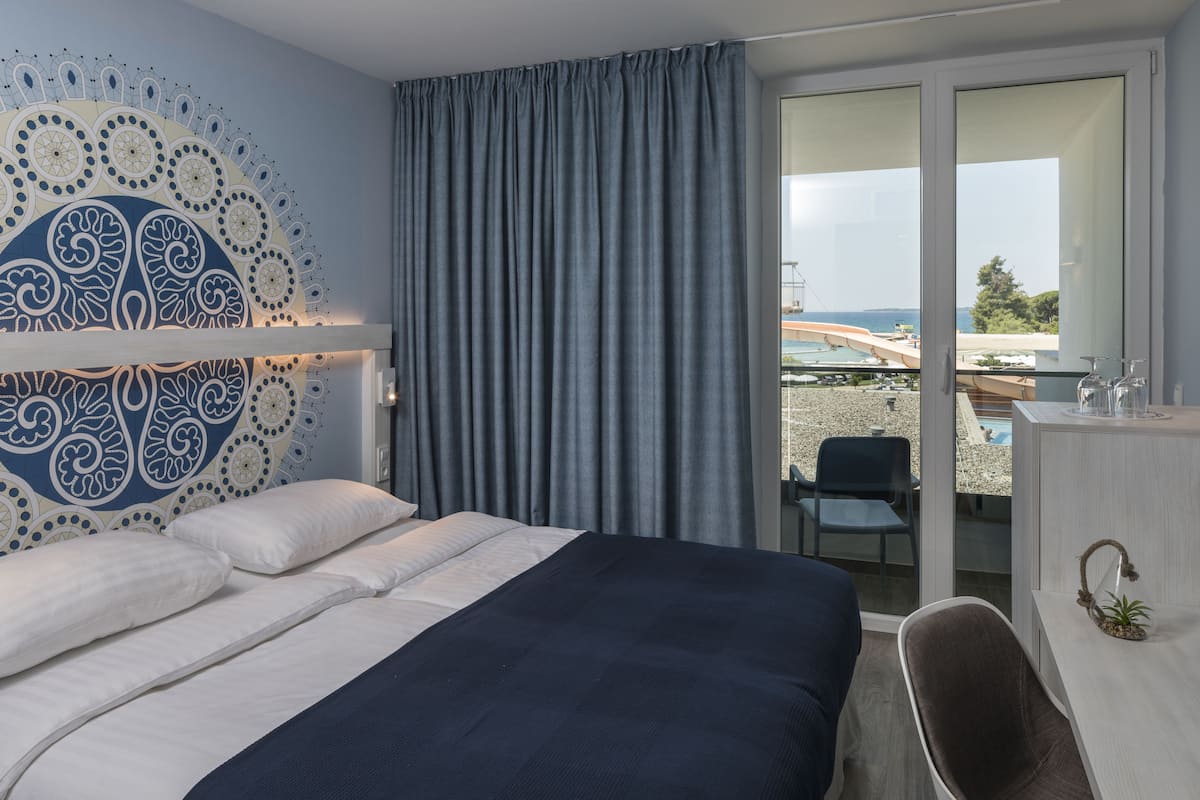 A modern hotel room with a large bed, a decorative wall, a sliding glass door to a balcony, and a small desk with a plant.
