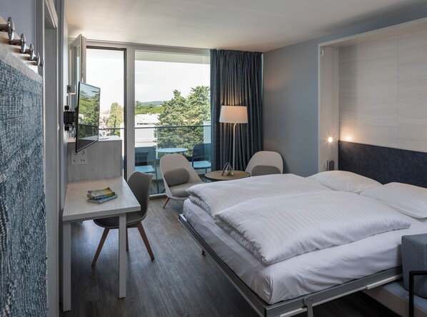 A hotel room with a bed, a desk, a chair, a small table, a lamp, and a view of the outdoors. - Falkensteiner Club Funimation Borik (Zadar)