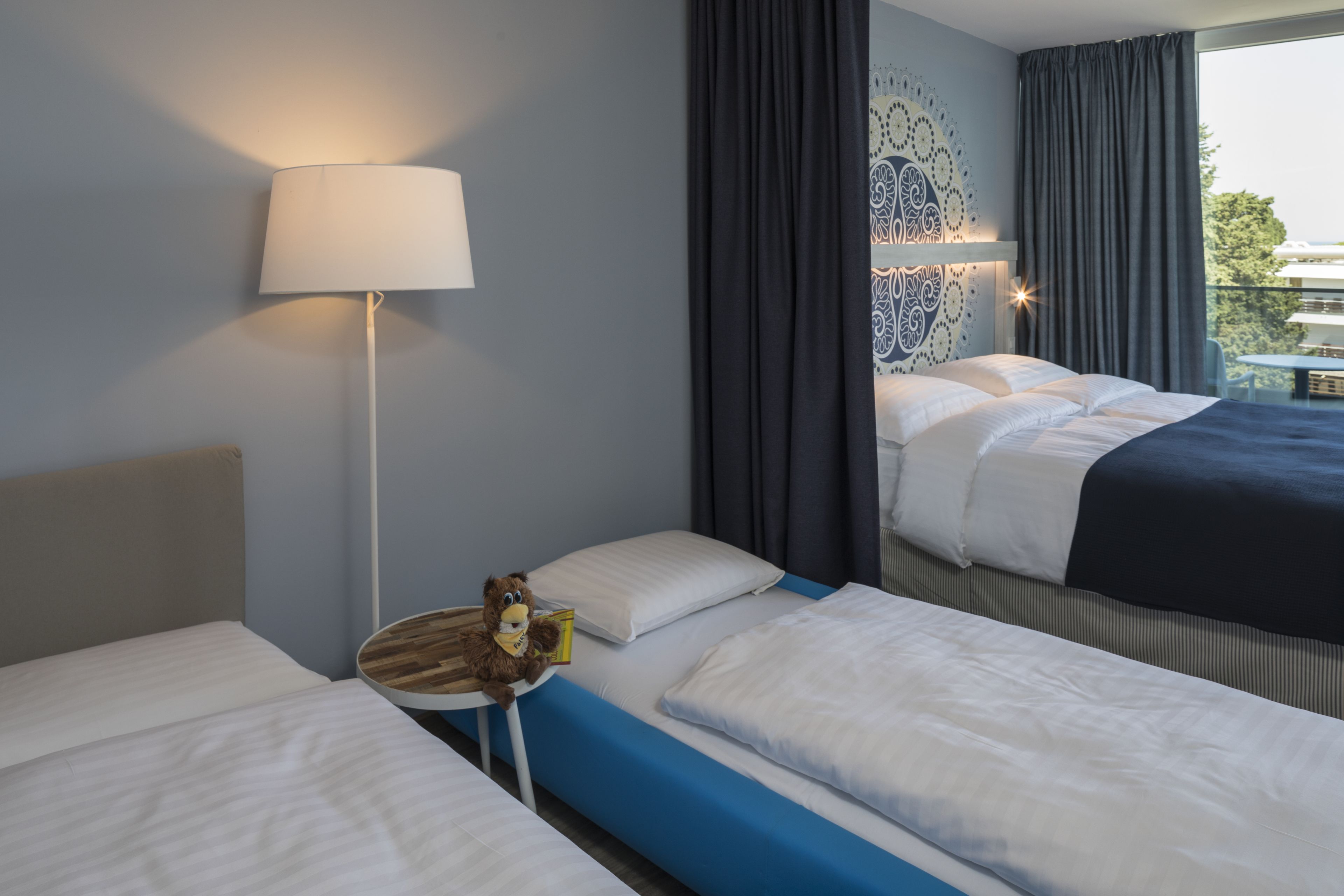 A hotel room with two beds, a lamp, and a view of the outside.