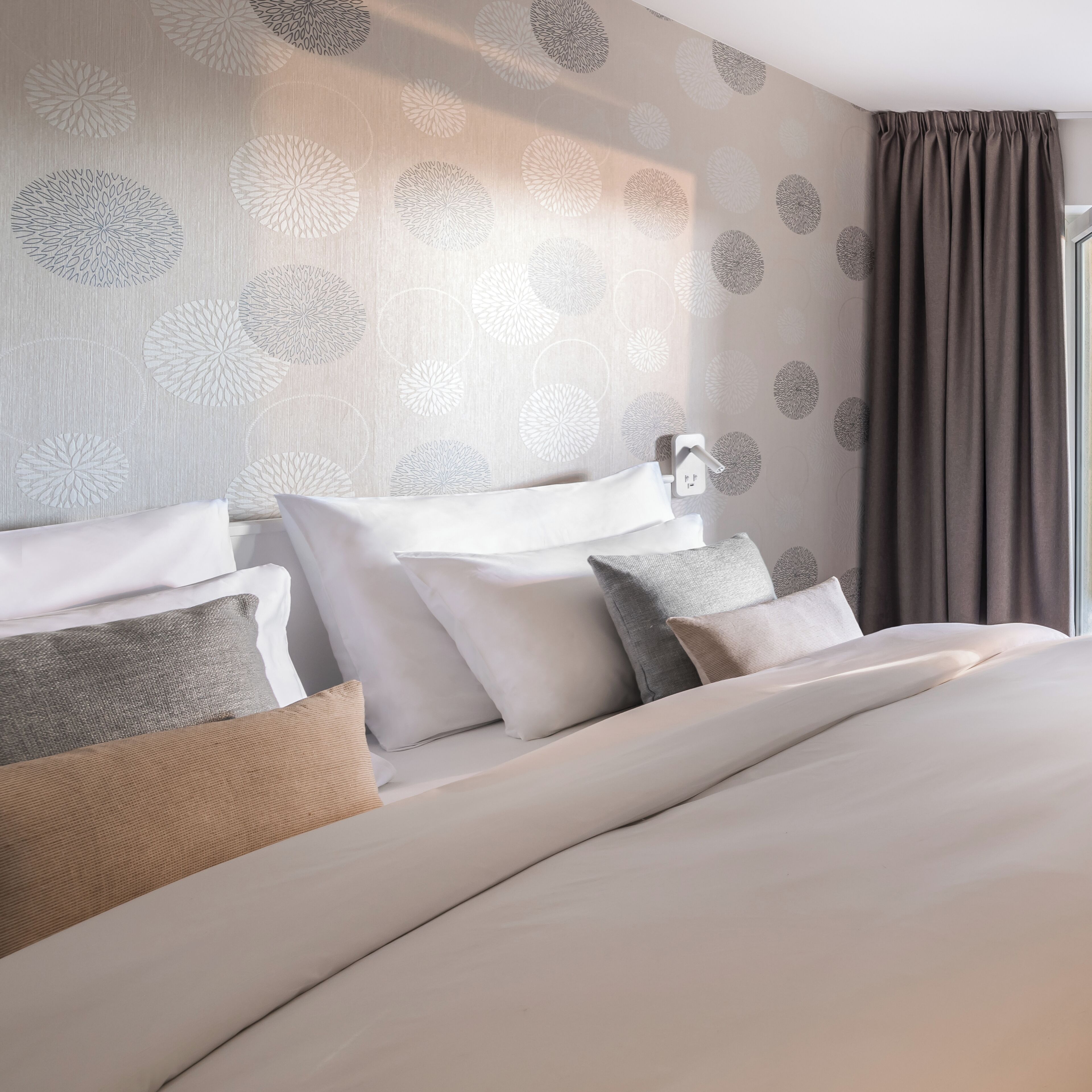 A neatly made bed with white and grey bedding, a wall with circular patterns, and a window with curtains.