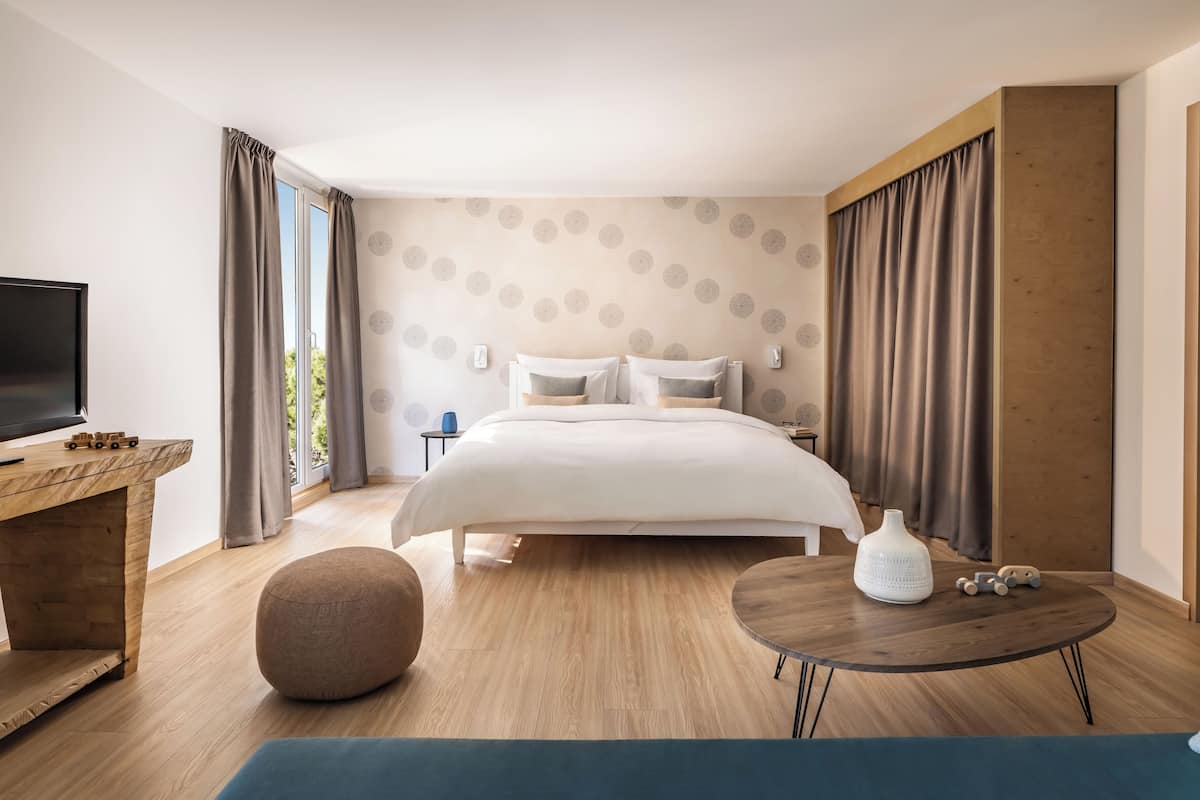 A modern hotel room with a large bed, a TV, a wooden desk, and a round table.