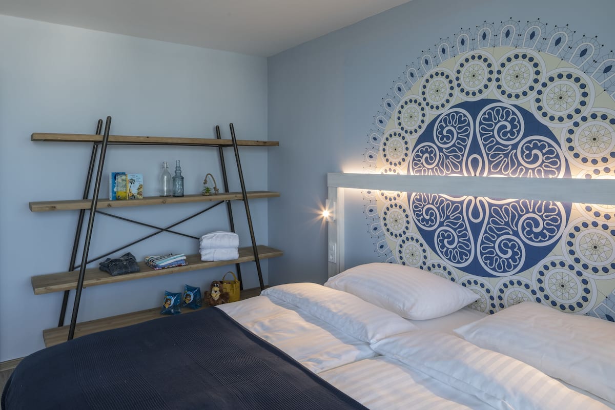 A bedroom with a bed, a headboard with a decorative pattern, wooden shelves with various items, and a wall tapestry.