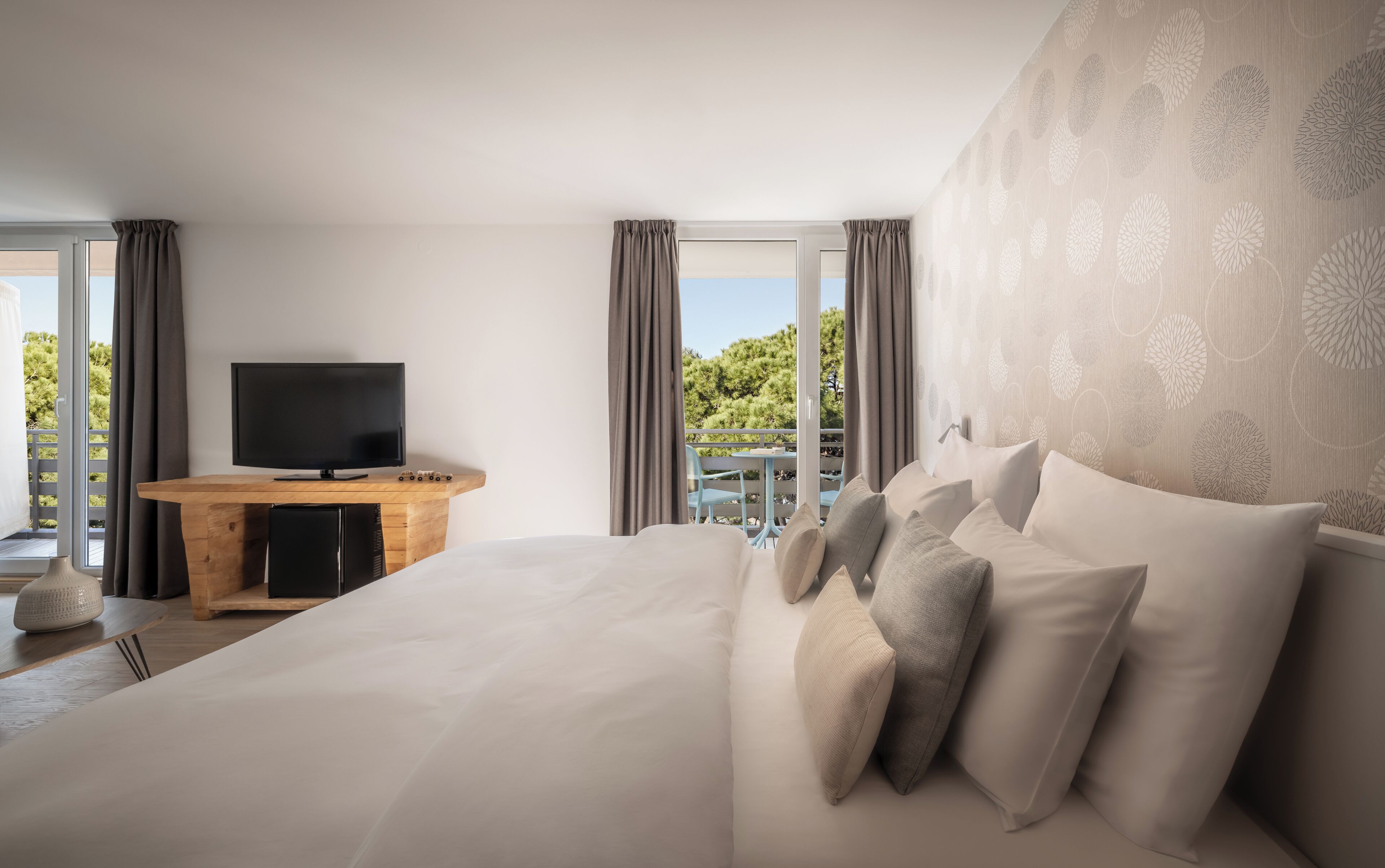 A bedroom with a large bed, a wooden desk with a TV, and a balcony with a table and chairs.