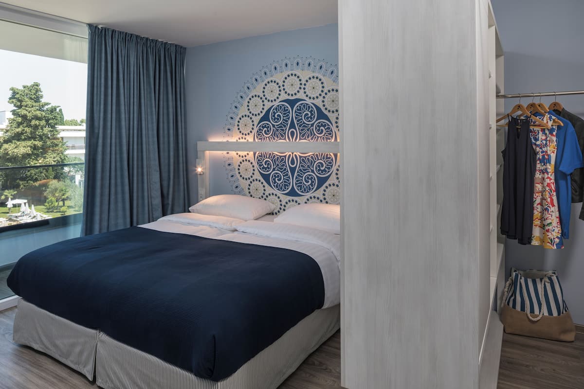 A modern bedroom with a large bed, a decorative headboard, a view of greenery outside, and a clothing rack with various garments.
