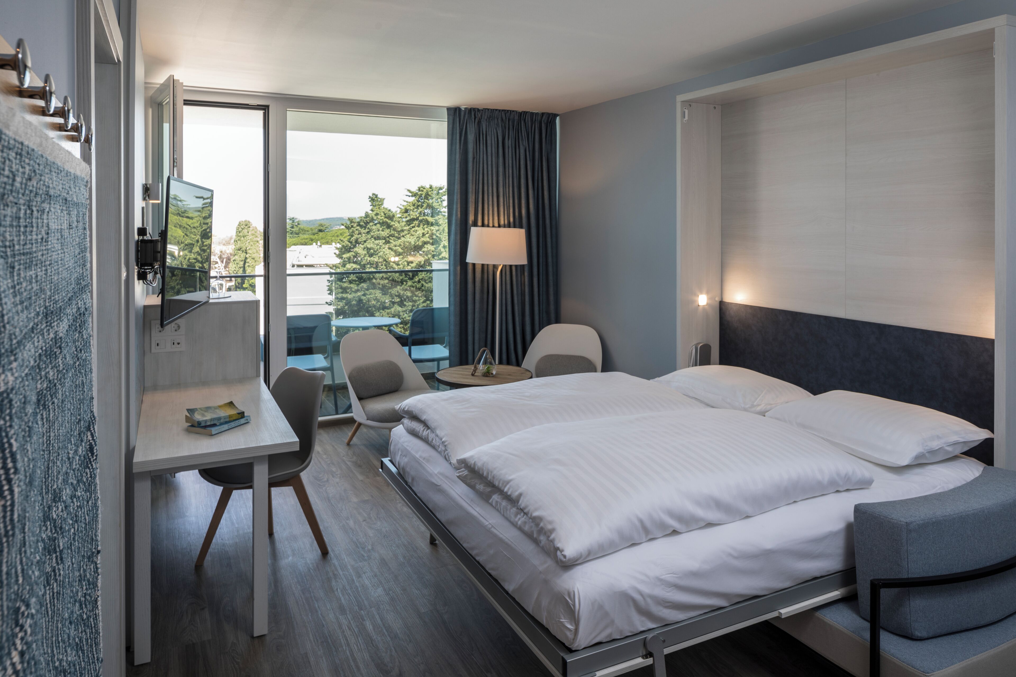 Ein Hotelzimmer mit Bett, Schreibtisch, Stühlen, einer Lampe und Blick ins Freie.