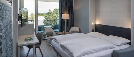 A hotel room with a bed, desk, chairs, a lamp, and a view of the outdoors.