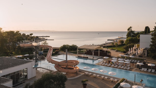 Zona de piscina en un resort con tobogán de agua, sillas de descanso, sombrillas y vista al mar.