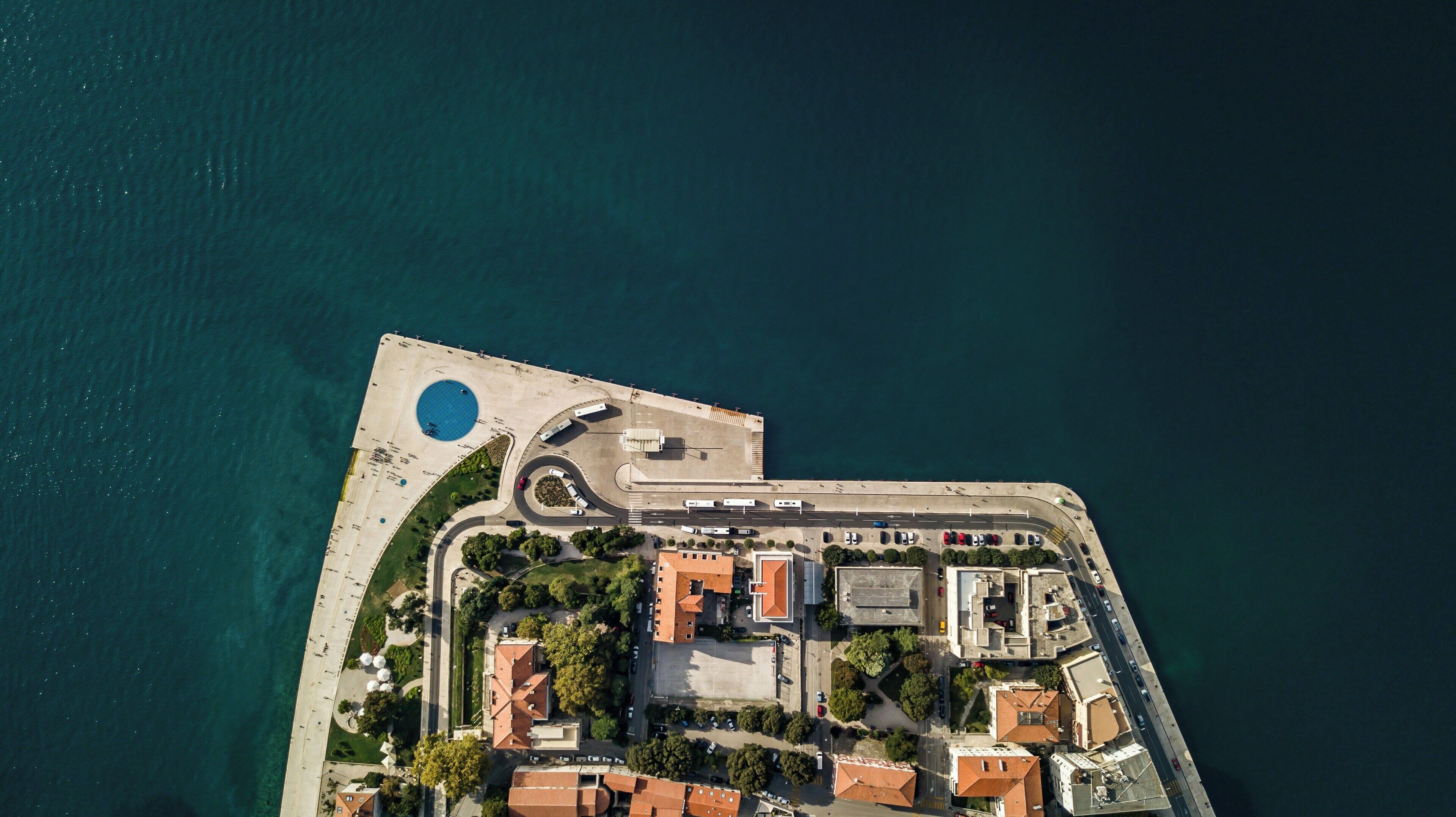 An aerial view of a coastal area with a marina, a roundabout, and buildings with red-tiled roofs.