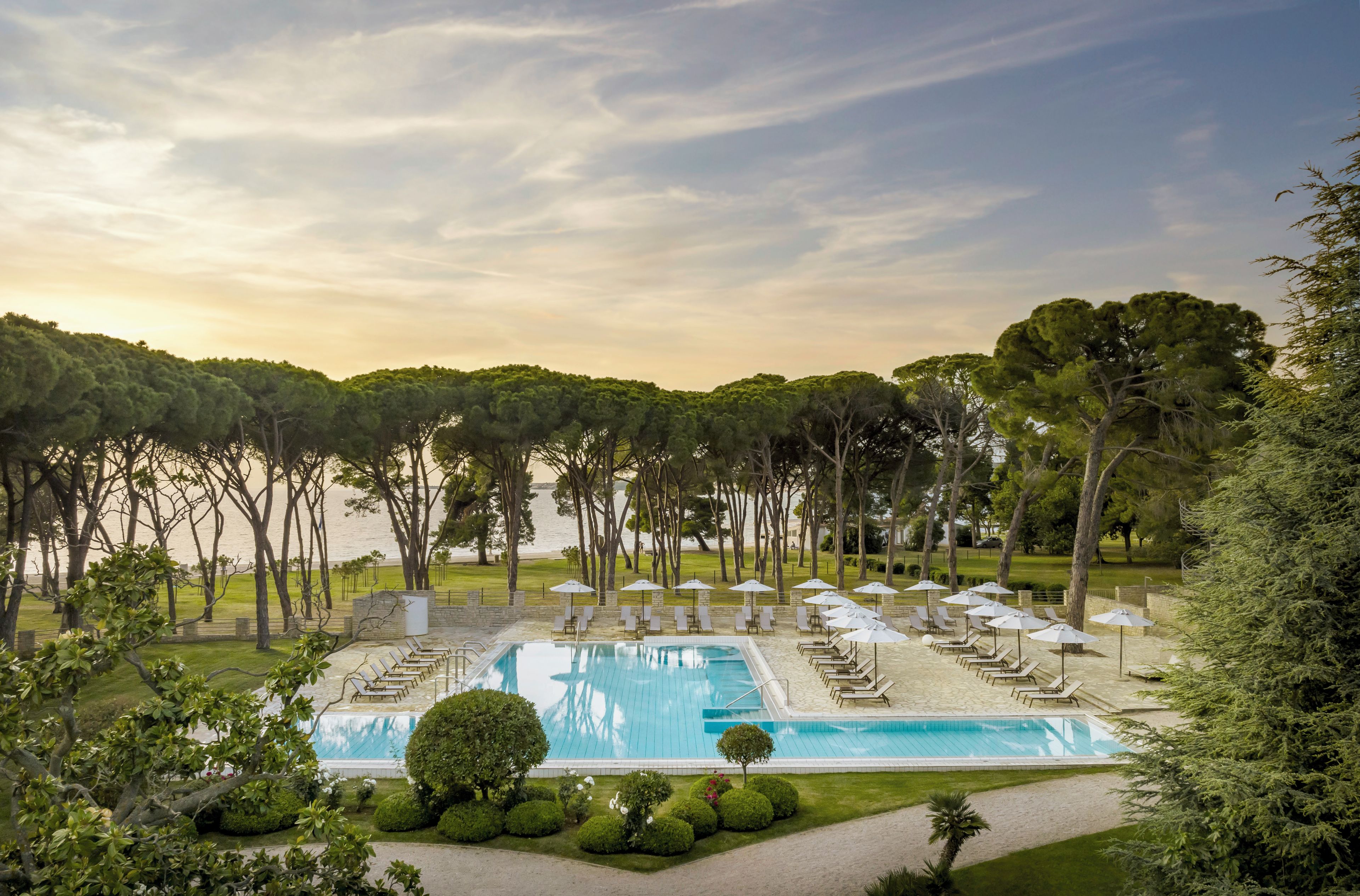 A pool area with lounge chairs and umbrellas, surrounded by trees and a view of the sea.