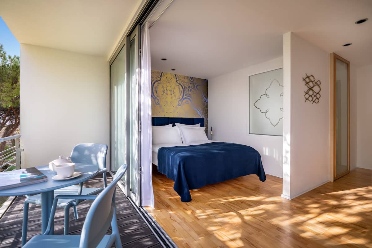 Un dormitorio con cama, una mesita de noche con taza y plato, una silla, y vistas al exterior.