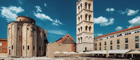 Una plaza histórica con una torre destacada y un edificio redondo, que ofrece mesas al aire libre y una fuente.