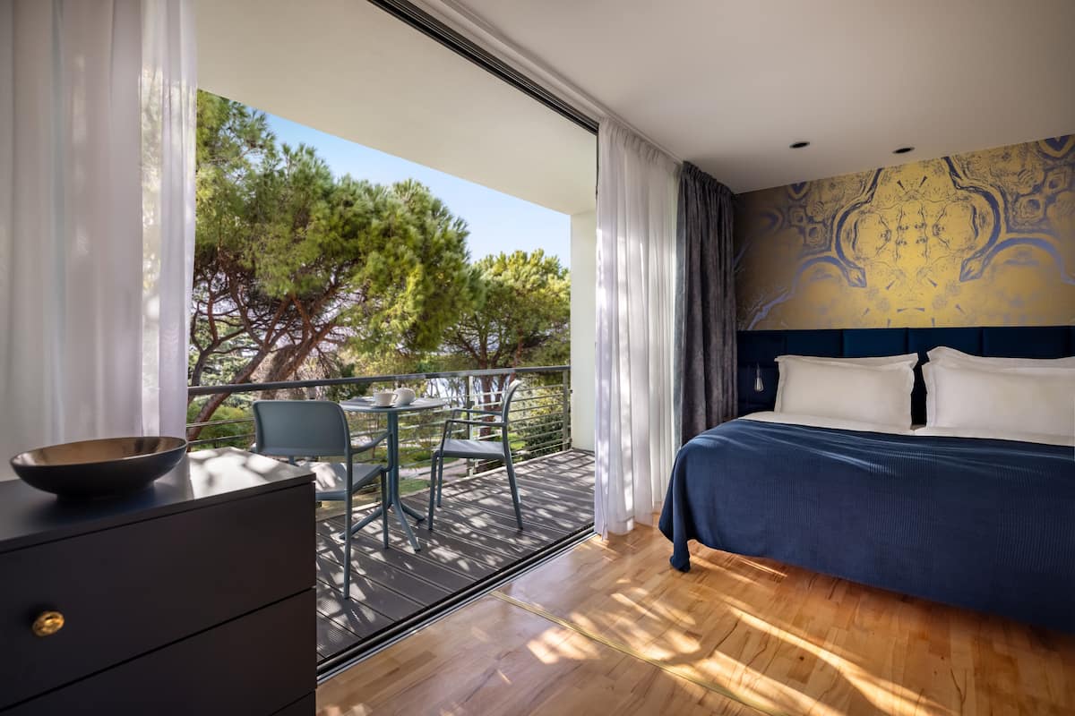 Un dormitorio con una cama grande, un aparador, una mesa con sillas y un balcón con vistas a árboles.