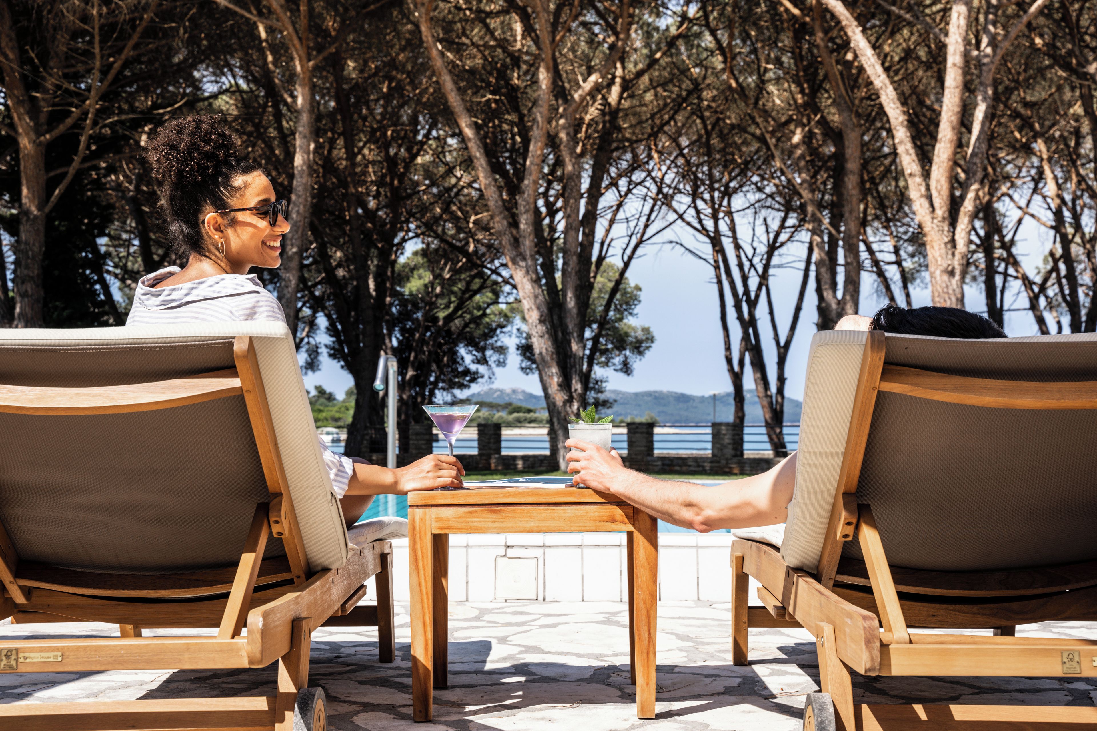 Une personne est assise à une table en bois, un verre à la main, et a devant elle une étendue d’eau et des arbres.