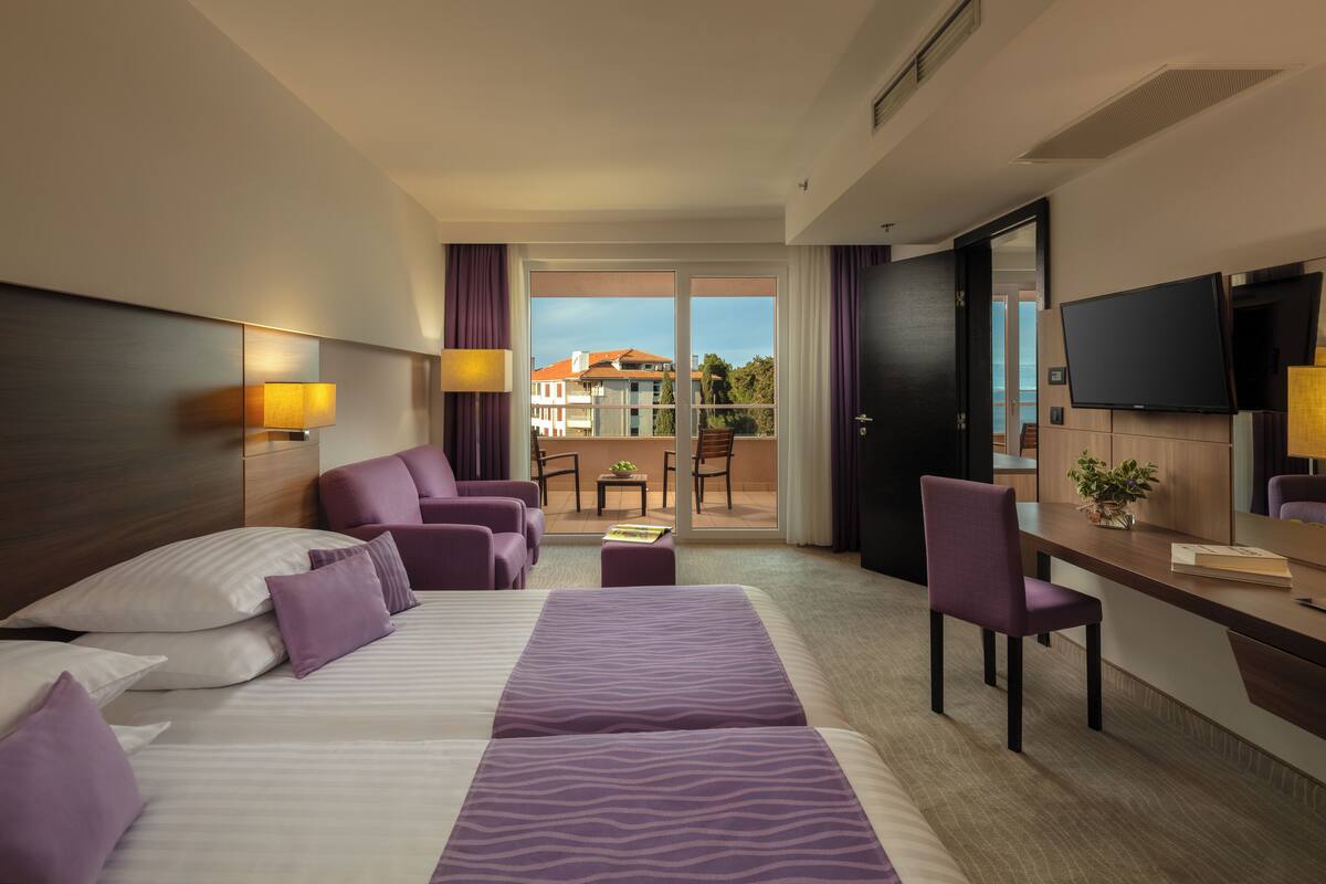 Habitación de hotel con una cama grande, un escritorio, una silla y un balcón con vistas a edificios.