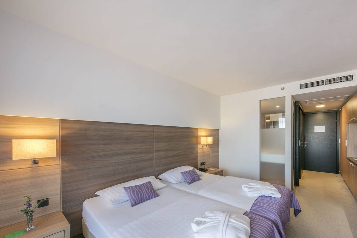 Habitación de hotel con dos camas, una mesita de noche, una lámpara, un espejo y una puerta que da a un baño.
