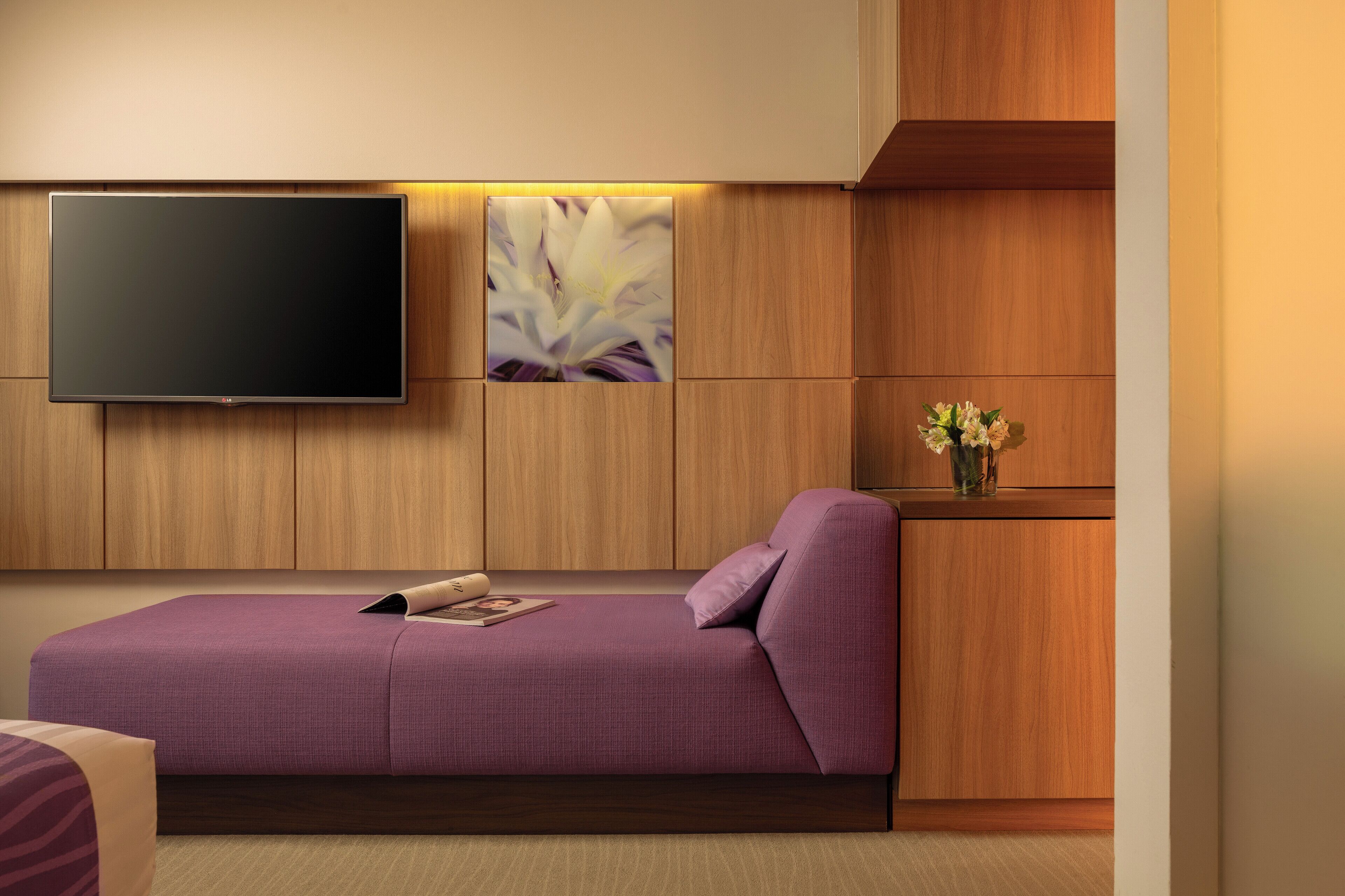 Une pièce moderne avec un canapé violet, un téléviseur à écran plat fixé au mur et un meuble en bois orné d’un vase de fleurs.