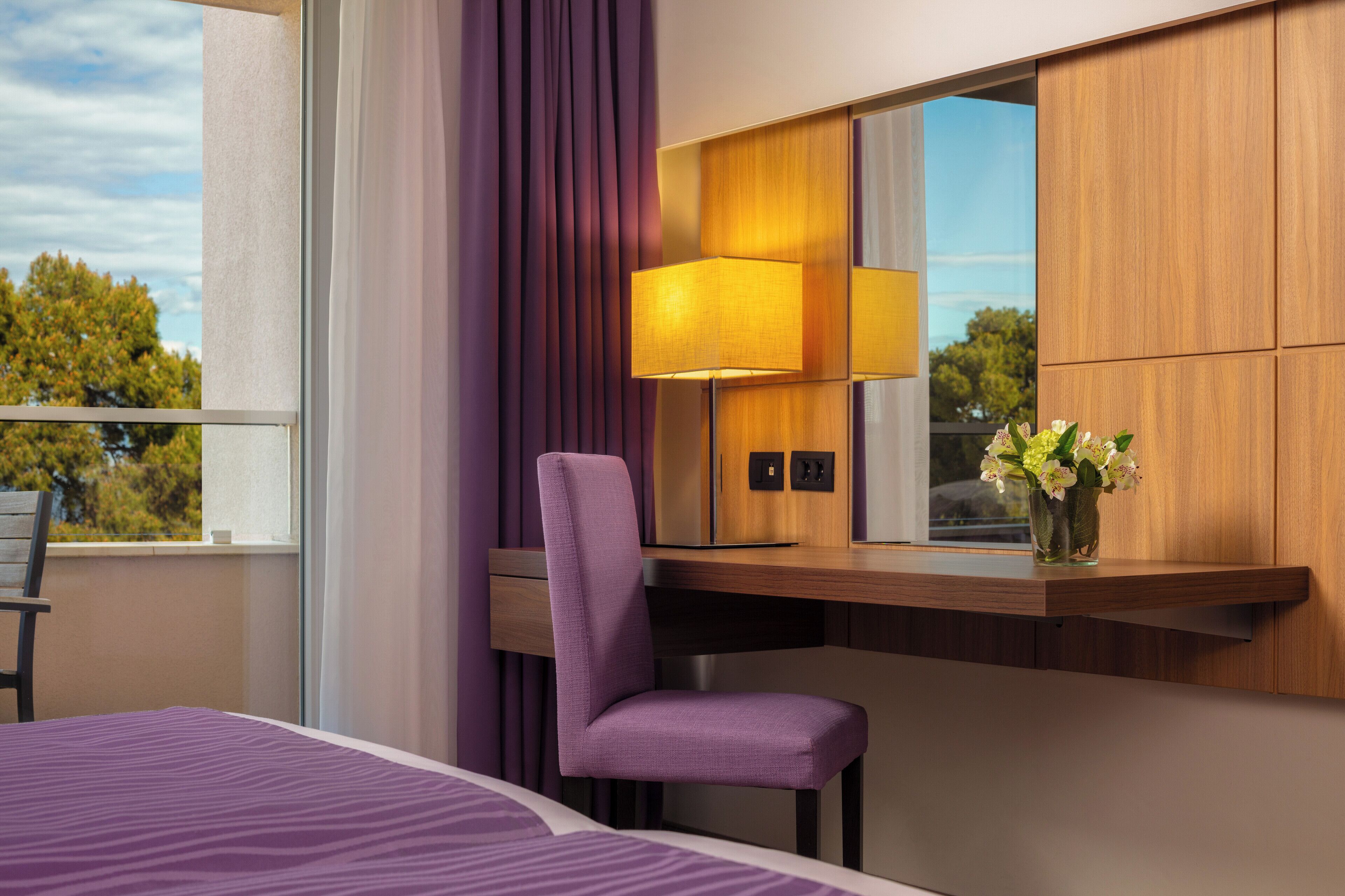 A hotel room with a bed, a desk with a lamp, a chair, a mirror, and a vase with flowers.
